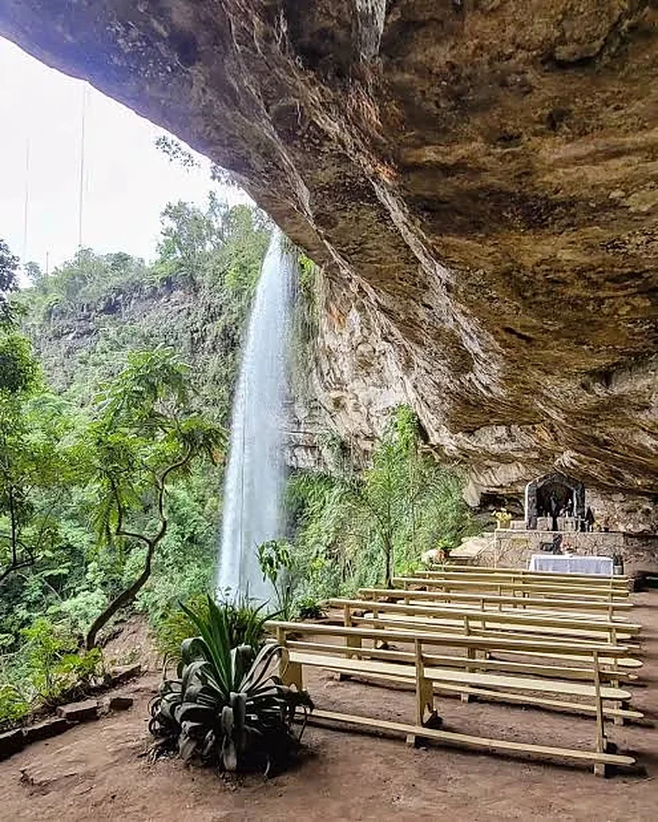 Cachoeira alta caindo ao lado da gruta do Poço Certo, com bancos e mata em volta
